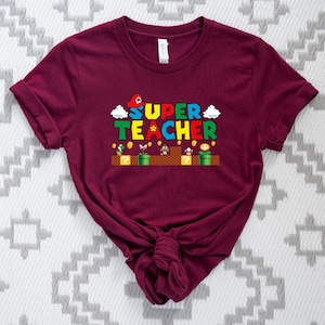 Puede incluir: Camiseta de manga corta color burdeos con un gr&aacute;fico de un personaje de videojuego y el texto "SUPER TEACHER".