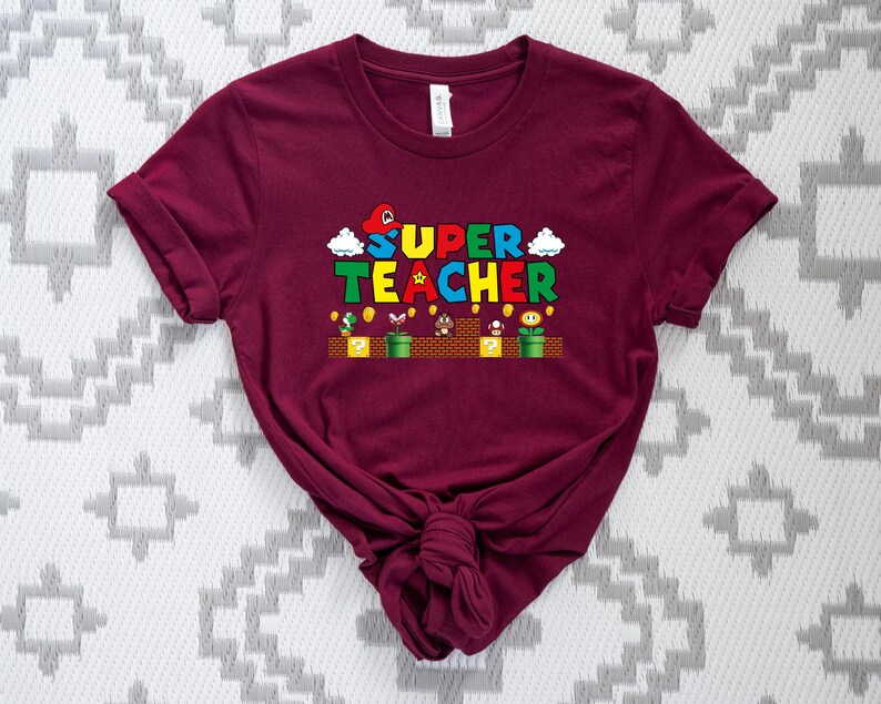 Puede incluir: Camiseta de manga corta color burdeos con un gr&aacute;fico de un personaje de videojuego y el texto "SUPER TEACHER".