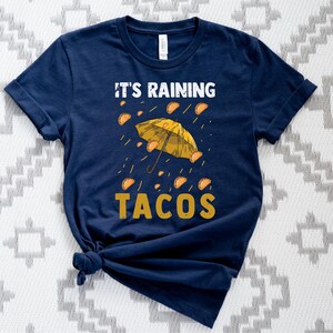 Peut inclure: Un t-shirt bleu marine avec un parapluie jaune et le texte "Il pleut des tacos".