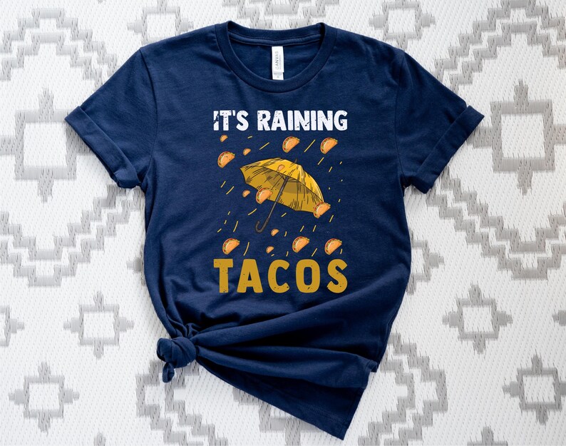 Peut inclure: Un t-shirt bleu marine avec un parapluie jaune et le texte "Il pleut des tacos".