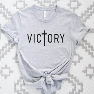 K&ouml;nnte beinhalten: Ein graues T-Shirt mit einem schwarzen Kreuz und dem Wort "VICTORY" auf der Vorderseite.