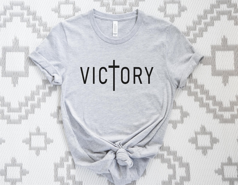 K&ouml;nnte beinhalten: Ein graues T-Shirt mit einem schwarzen Kreuz und dem Wort "VICTORY" auf der Vorderseite.