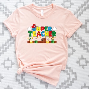 Puede incluir: Una camiseta rosa con un gr&aacute;fico tem&aacute;tico de Super Mario Bros. que dice "SUPER TEACHER" con un hongo, una nube y un bloque de signo de interrogaci&oacute;n.