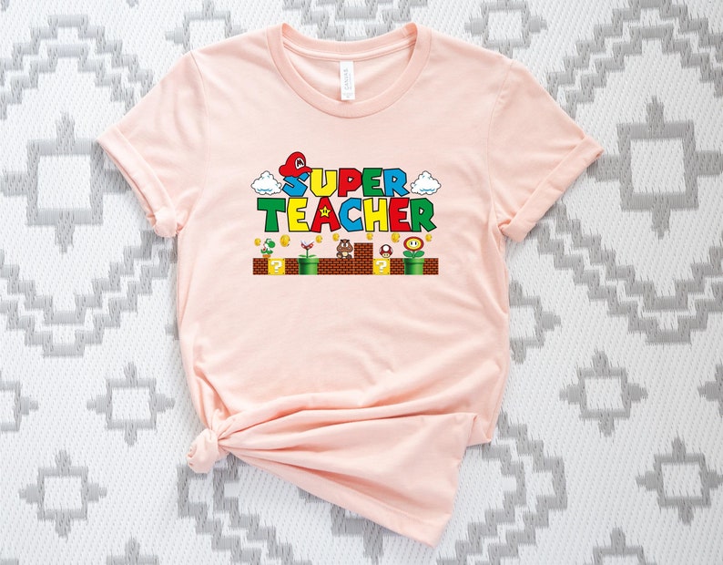 Puede incluir: Una camiseta rosa con un gr&aacute;fico tem&aacute;tico de Super Mario Bros. que dice "SUPER TEACHER" con un hongo, una nube y un bloque de signo de interrogaci&oacute;n.