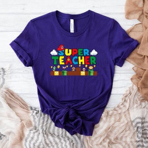 Puede incluir: Camiseta morada con un gr&aacute;fico de un personaje de videojuego y el texto "SUPER TEACHER".