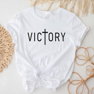 K&ouml;nnte beinhalten: Wei&szlig;es T-Shirt mit einem schwarzen Kreuz und dem Wort "VICTORY" auf der Vorderseite.
