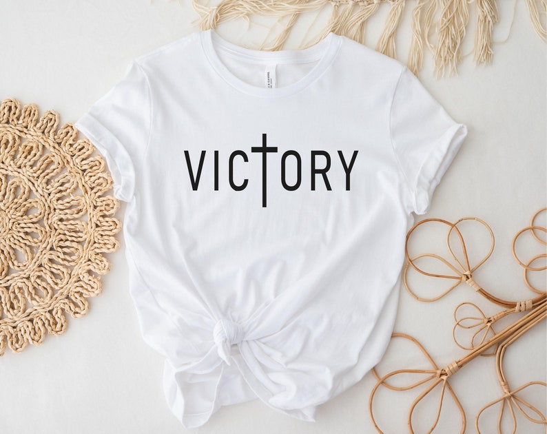 K&ouml;nnte beinhalten: Wei&szlig;es T-Shirt mit einem schwarzen Kreuz und dem Wort "VICTORY" auf der Vorderseite.