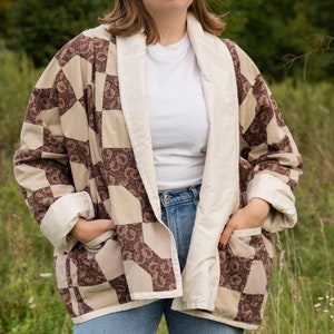 May include: A woman wearing a brown and white quilted jacket with a white collar and a white t-shirt underneath. She is wearing blue jeans and standing in a field of tall grass.