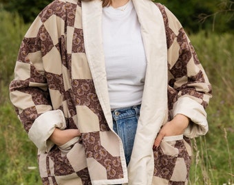 Può includere: Una donna indossa una giacca trapuntata marrone e bianca con un colletto bianco e una maglietta bianca sotto. Indossa jeans blu e si trova in un campo di erba alta.