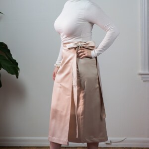 May include: A woman wearing a white turtleneck sweater and a blush pink wrap skirt. She is wearing brown suede ankle boots with buckles.
