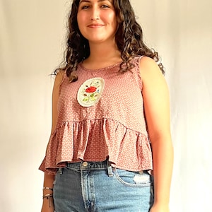 May include: A pink sleeveless top with a white floral embroidered design. The top has a ruffled hem and a fitted bodice. The model is wearing blue jeans.