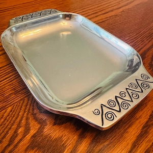 May include: A rectangular silver-toned metal serving tray with a smooth, reflective center surface. The two opposite handles feature a decorative black etched pattern consisting of repeating zigzag lines and small spiral motifs at each point.