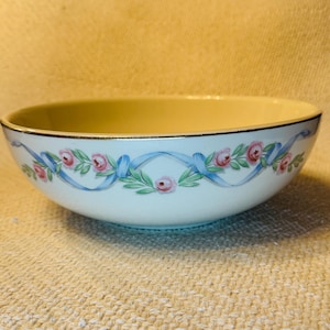 May include: A white ceramic bowl with a cream-colored interior and a gold rim. The exterior features a decorative pattern of pink roses, green leaves, and blue ribbons. The bowl is likely for serving food.