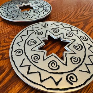 May include: Two circular silver-toned metal trivets featuring a central eight-pointed star cutout. The surface is decorated with raised black painted patterns, including spiral motifs surrounding the star and a zigzag border along the outer edge.