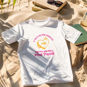 May include: White t-shirt with a graphic design. The design features a heart with the word "ME" inside, surrounded by text that reads "If ten is the main character, I'm his perfect plot twist."