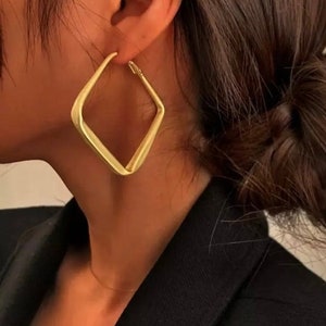May include: A woman with dark brown hair styled in a bun is wearing large, gold, square-shaped hoop earrings.