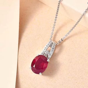 May include: A silver chain necklace with a pendant featuring a large oval-shaped ruby gemstone surrounded by smaller clear gemstones.