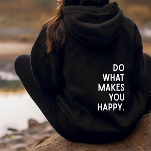 Könnte beinhalten: Schwarzer Kapuzenpullover mit dem weißen Text "DO WHAT MAKES YOU HAPPY". Der Kapuzenpullover wird von einer Person im Freien getragen. Der Hintergrund ist verschwommen und zeigt ein Gewässer und eine natürliche Umgebung.