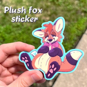 May include: A die-cut sticker featuring a cartoon plush fox with orange and cream fur, dark purple paws, and a pink bow tie around its neck. The fox has a stitched appearance, large ears, and a long tail with a dark purple band near the tip.