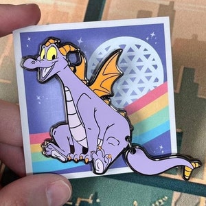 May include: Purple and orange enamel pin featuring a cartoon dragon with a rainbow and a blue sphere with a geometric pattern in the background.