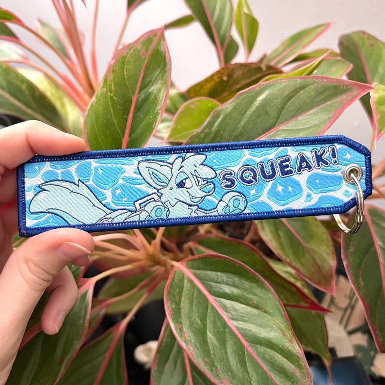 May include: A light blue embroidered keychain with a cartoon fox swimming in water. The keychain has the word "SQUEAK!" in white text and a silver key ring. The background is a plant with green and pink leaves.