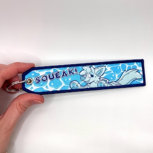 May include: A blue and white embroidered keychain with a cartoon fox character and the word "SQUEAK!". The keychain has a blue border and a silver key ring.