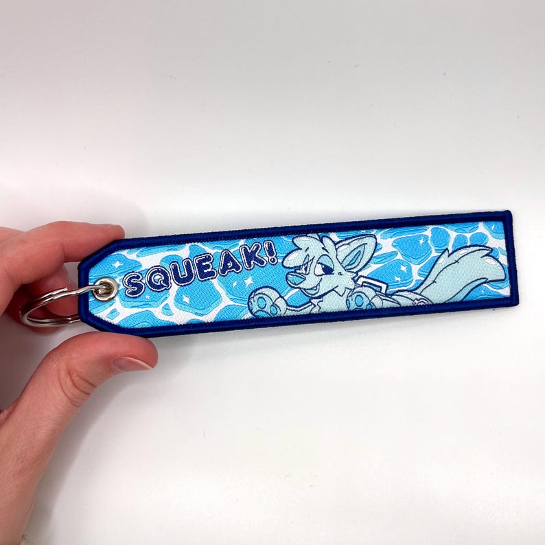 May include: A blue and white embroidered keychain with a cartoon fox character and the word "SQUEAK!". The keychain has a blue border and a silver key ring.