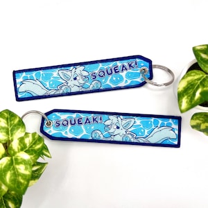 May include: Two embroidered keychains with a light blue and white cartoon fox design. The keychains have a blue border and the word "SQUEAK!" in white. Each keychain has a silver keyring.