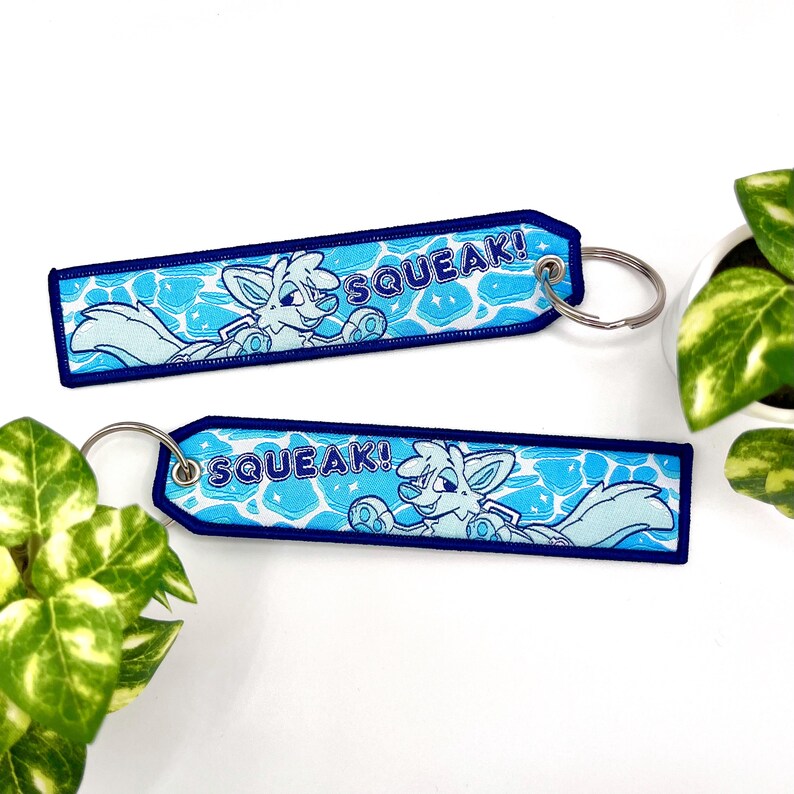 May include: Two embroidered keychains with a light blue and white cartoon fox design. The keychains have a blue border and the word "SQUEAK!" in white. Each keychain has a silver keyring.