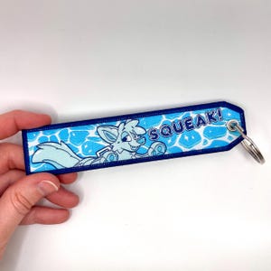 May include: A blue and white embroidered keychain with a cartoon fox illustration and the word "SQUEAK!". The keychain has a blue border and a silver key ring.