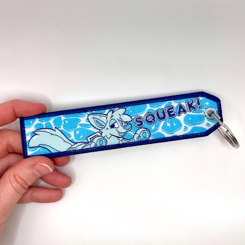 May include: A blue and white embroidered keychain with a cartoon fox illustration and the word "SQUEAK!". The keychain has a blue border and a silver key ring.
