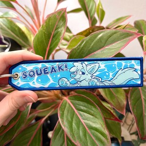 May include: A blue and white embroidered keychain with the word "SQUEAK!" and a cartoon fox character. The keychain has a blue border and a silver key ring. The background features a water-like design.