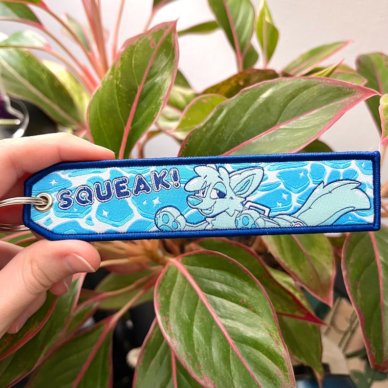 May include: A blue and white embroidered keychain with the word "SQUEAK!" and a cartoon fox character. The keychain has a blue border and a silver key ring. The background features a water-like design.