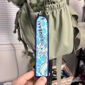 May include: A rectangular keychain with a blue border and a cartoon fox illustration. The keychain has the word "SQUEAK!" in white text. The background features a blue water pattern. The keychain is attached to a silver keyring.