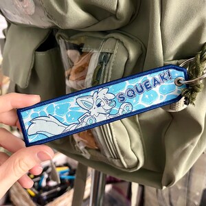 May include: A blue and white embroidered tag with the word "SQUEAK!" and a cartoon fox character. The tag is attached to a green backpack. The fox is swimming in a pool of water.