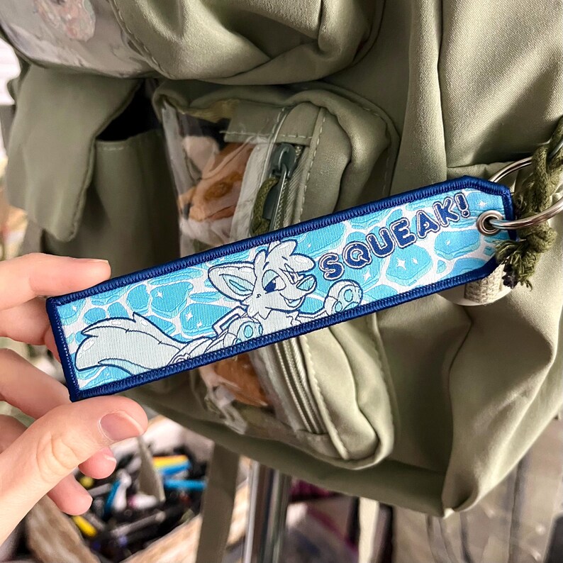 May include: A blue and white embroidered tag with the word "SQUEAK!" and a cartoon fox character. The tag is attached to a green backpack. The fox is swimming in a pool of water.