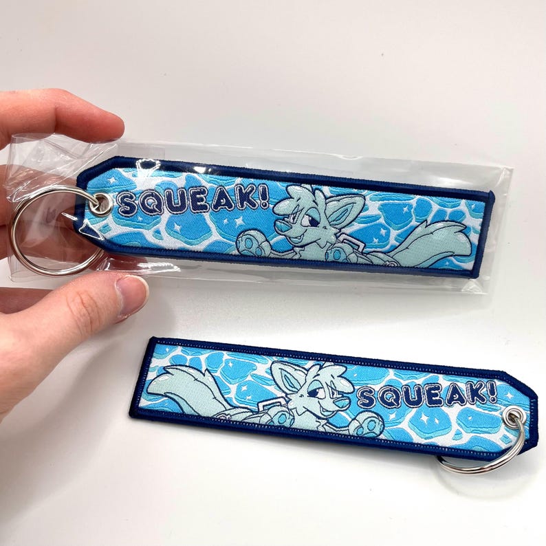 May include: Two blue and white embroidered keychains with a cartoon fox design and the word "SQUEAK!". The keychains have a blue border and a silver key ring attached. The background features a water-like pattern.