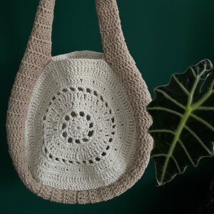 May include: A handmade crochet bag in cream and tan. The bag features a circular design with a spiral pattern in the center. The strap and outer edge are tan, while the interior is cream-colored. The bag is displayed against a dark green background.