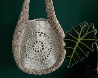 May include: A handmade crochet bag in cream and tan. The bag features a circular design with a spiral pattern in the center. The strap and outer edge are tan, while the interior is cream-colored. The bag is displayed against a dark green background.
