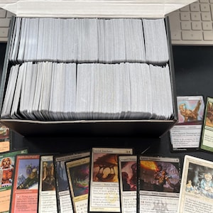 May include: A large black cardboard storage box filled with rows of white trading cards, positioned in front of a computer keyboard. Several individual trading cards with colorful illustrations and printed text are displayed in front of the box.