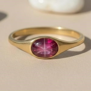 Peut inclure: Une bague en or avec une pierre précieuse ovale en rubis en forme d'étoile. La bague est posée sur une surface beige clair, avec plusieurs pierres blanches et arrondies en arrière-plan. Le rubis est d'un rouge profond.