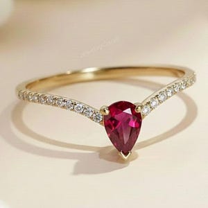 May include: A gold ring featuring a pear-shaped, deep red gemstone. The band is embellished with small, clear gemstones. The ring has a V-shaped design, and the gemstones are set in a delicate style. The ring is displayed on a light-coloured surface.