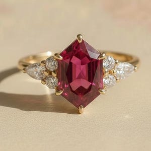Può includere: Un anello d'oro con una grande pietra preziosa rosso-rosa sfaccettata a forma esagonale. L'anello è impreziosito da pietre preziose più piccole e trasparenti su entrambi i lati della pietra principale. La fascia dell'anello è in oro lucido.