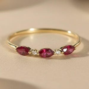 Op de afbeelding: Een delicate gouden ring met drie marquise-geslepen robijnen edelstenen en twee kleine diamantaccenten. De ring heeft een dunne band en een gepolijste afwerking, die het levendige rood van de robijnen benadrukt.