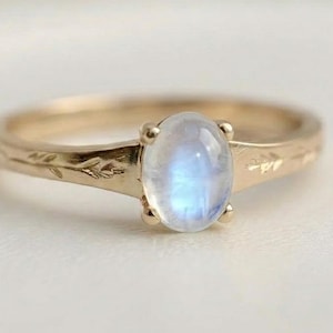 Op de afbeelding: Een gouden ring met een ovale, lichtblauwe edelsteen. De band heeft een delicaat bloemmotief. De ring staat tegen een zachte, neutrale achtergrond, waardoor de kleur van de edelsteen en de details van de ring worden benadrukt.