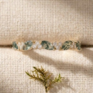 May include: A gold band ring featuring alternating marquise-cut moss agate stones and small round clear stones. The ring has a delicate, nature-inspired design with green inclusions visible within the translucent moss agate gemstones.