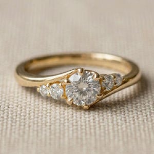 Può includere: Un anello d'oro con una grande gemma rotonda e trasparente al centro, affiancata da gemme più piccole e trasparenti. L'anello ha un design curvo, con le gemme incastonate in un'incastonatura a griffe. L'anello è su una superficie di colore neutro.