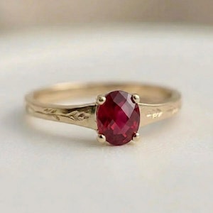 Puede incluir: Un anillo de oro con una piedra preciosa de color rojo intenso, talla ovalada y facetada, montada en un engaste de cuatro garras. La banda es de oro amarillo y presenta un delicado patrón de hojas grabado en los laterales.