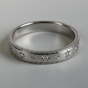 May include: A silver ring with a star pattern. The ring features a band of small, raised dots along the edges and a series of engraved stars, some with small, clear stones. The ring is set against a neutral background.