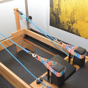 May include: A wooden Pilates reformer with blue straps and a black padded seat. The straps are attached to pulleys and have colorful patterned bands.